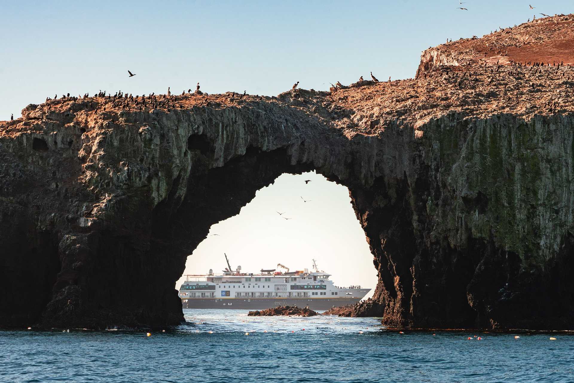 a large rock arch frames a ship. birds are perched on top of the rocks.