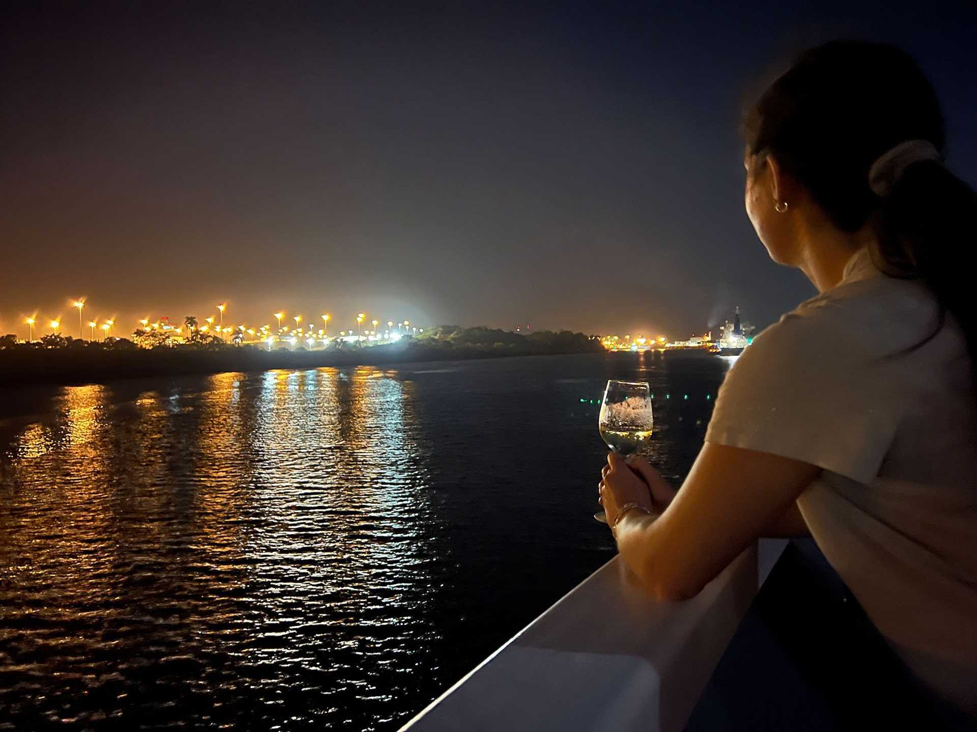 a woman stands on the deck of a ship holding wine and watching the panama canal at night