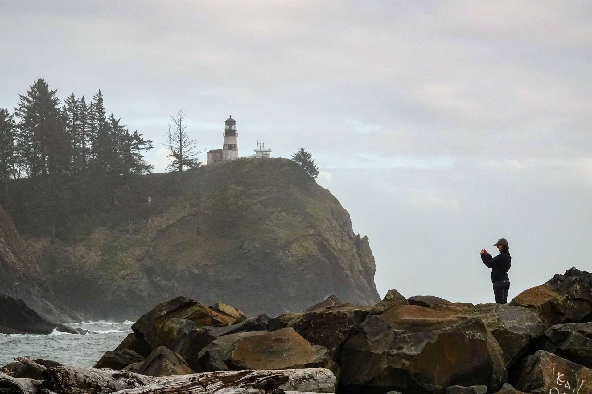 a woman stands on rocks and photographs a distant lighthouse
