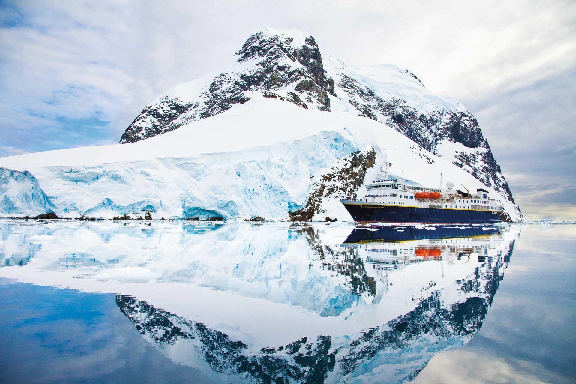 The National Geographic Explorer sails through Lemaire Channel with snowy mountains and a glacier behind it.