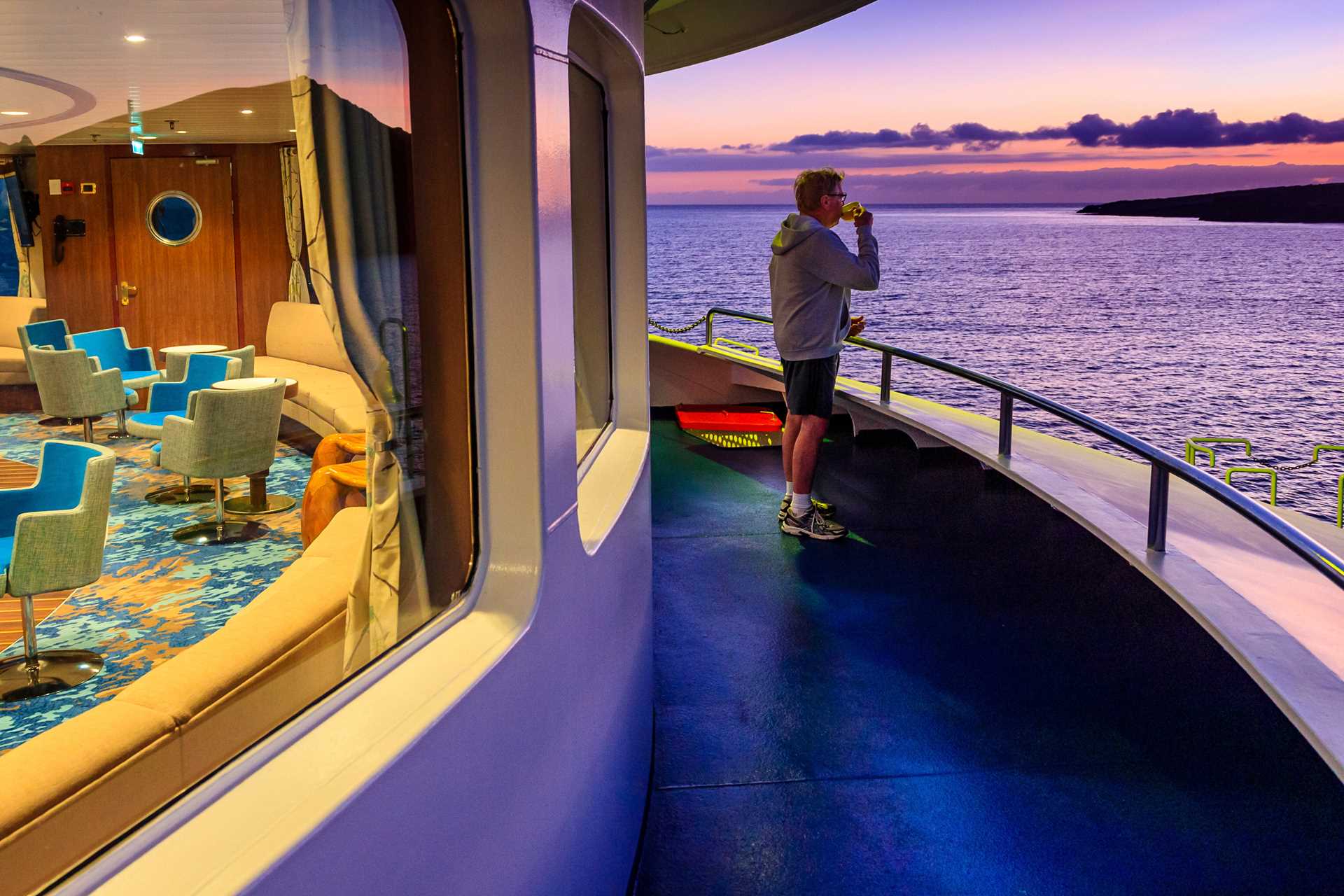 A guest onboard on deck looks out into the horizon during twilight while having a drink.