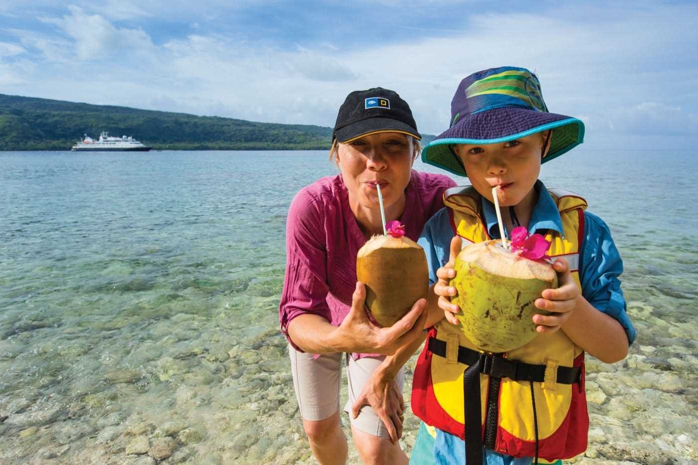 Two people—and adult and a child—drink out of coconuts using straws. In the background, a cruise ship sits in the bay