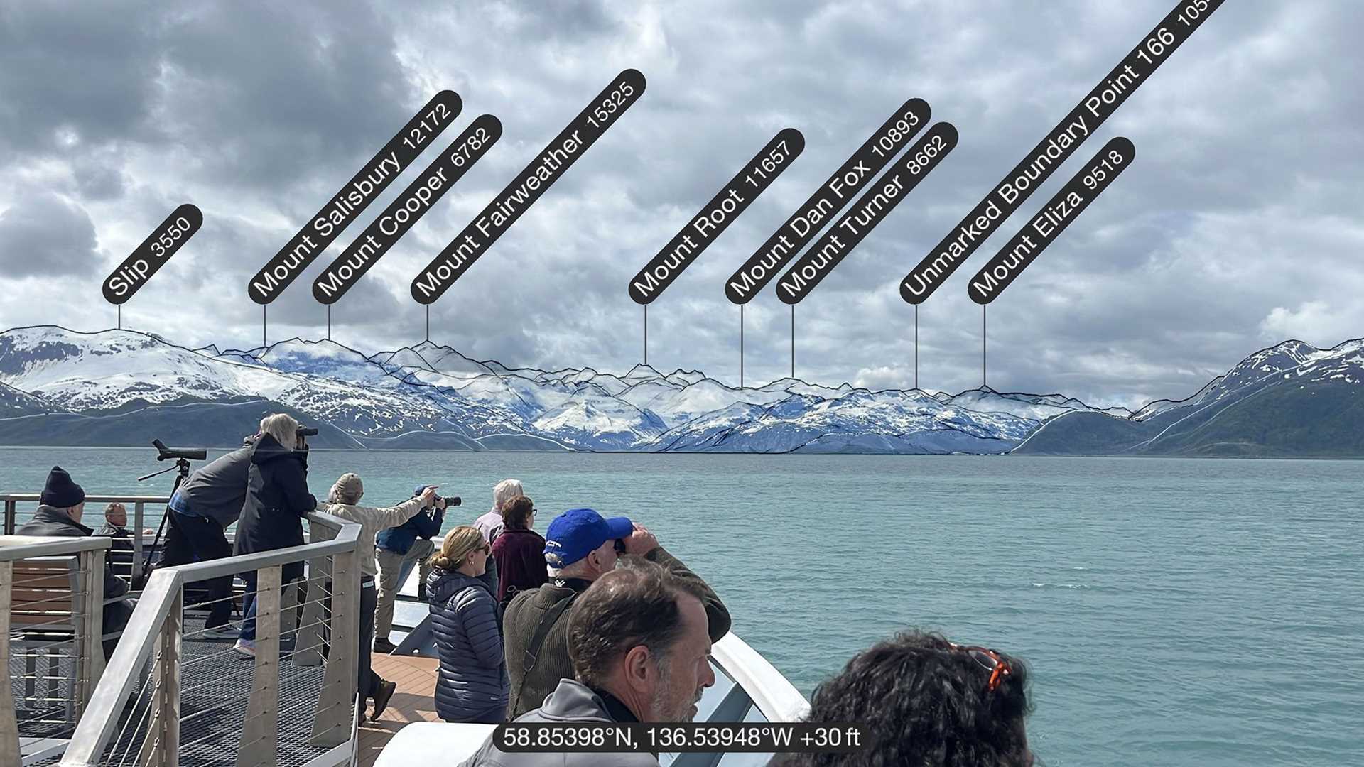 guests on the deck of a ship with labeled mountain peaks in the background