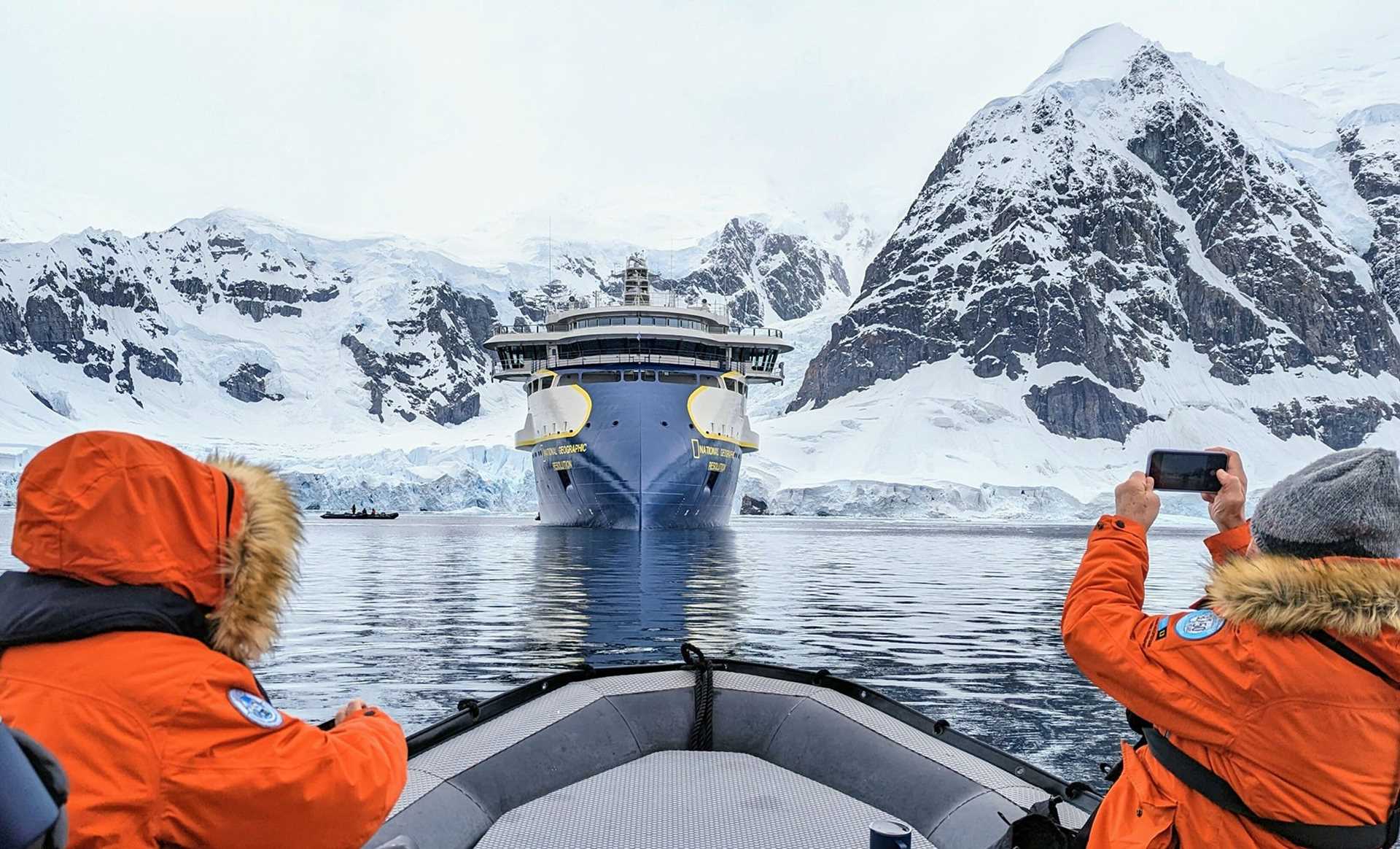 people in orange parkas in a zodiac, observing glaciers and a ship